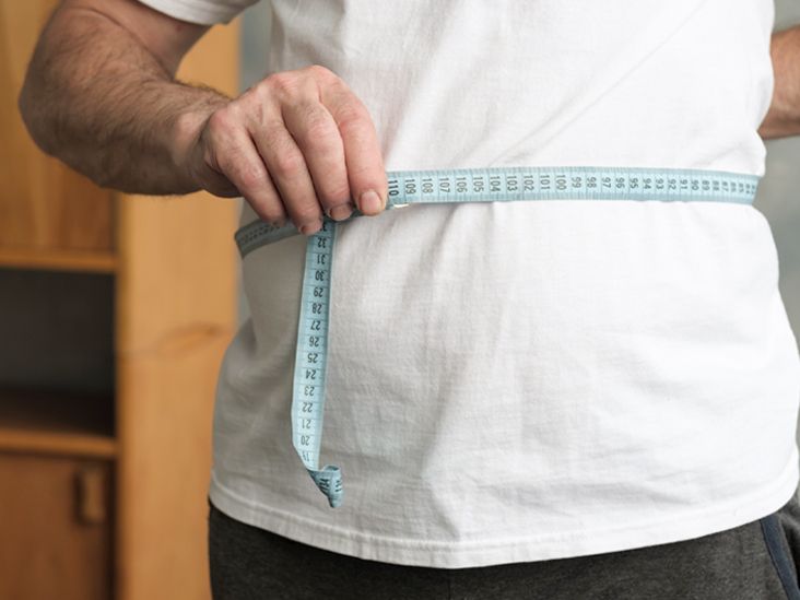man measuring his belly with measurement tape standing in the living room.